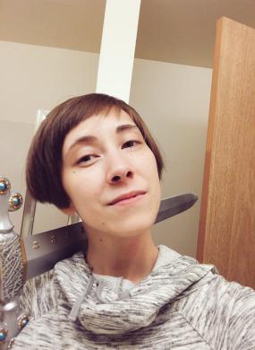 Headshot of Kiku Hughes (Kiku has short brown hair and is looking into the camera, she is holding a sword behind her head)